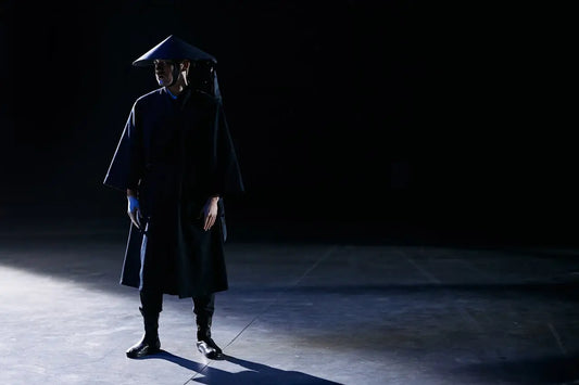 A solemn samurai man dressed in all black with a traditional straw kasa hat, standing straight indoors with his head turned to the side, expressing indifference and self-mastery