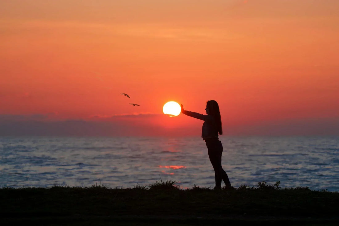 A young woman by the ocean at sunset, raising her arm as if to hold the sun, a symbol of embracing the here and now